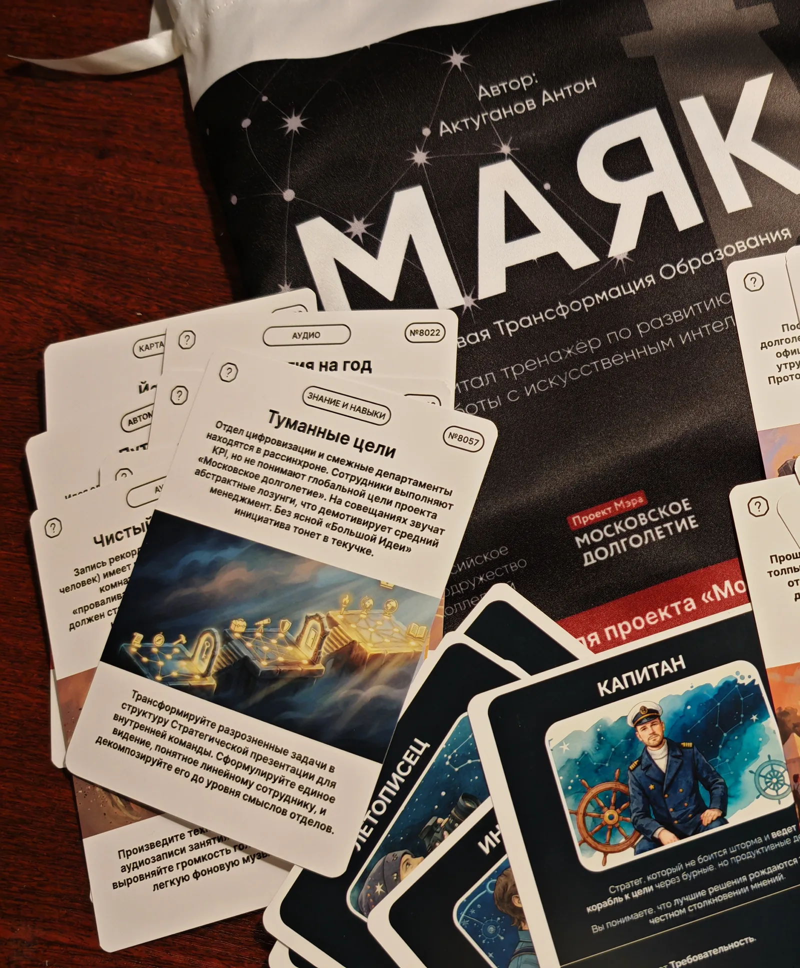 Чехол комплекта «МАЯК» с именем автора на лицевой стороне, рядом веером разложены карточки заданий и тёмные ролевые карты